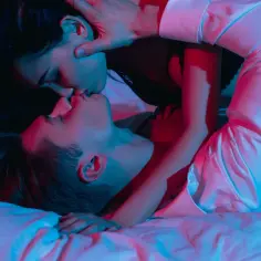 Para zbliża się do siebie w intymnym uścisku, tworząc ciekawe pozycje seksualne. Czerwone i niebieskie światła podkreślają ich bliskość.