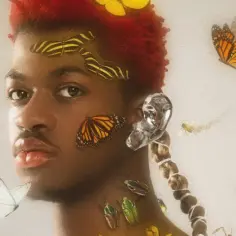 Lil Nas X, otoczony motylami i ważkami, z czerwonymi włosami i srebrnym kolczykiem. Czy Lil Nas X jest gejem? To pytanie nurtuje fanów.