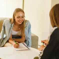 Uśmiechnięta kobieta w okularach pisze coś w notatniku. Rozmowa z terapeutą, czyli psychoterapia co to jest, może pomóc w zrozumieniu siebie.