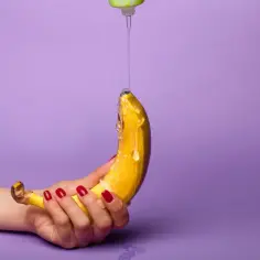 Dłoń z czerwonymi paznokciami trzyma banana, na który kapie przezroczysty lubrykant. Jaki lubrykant do prezerwatywy?