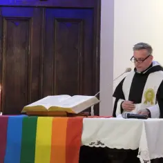 Ksiądz gej w szatach liturgicznych, z tęczową flagą i świecami, przemawia przy mikrofonie.