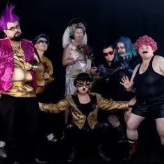 Grupa drag kingów w ekstrawaganckich strojach, z kolorowymi perukami i błyszczącymi dodatkami, pozuje do zdjęcia na czarnym tle.